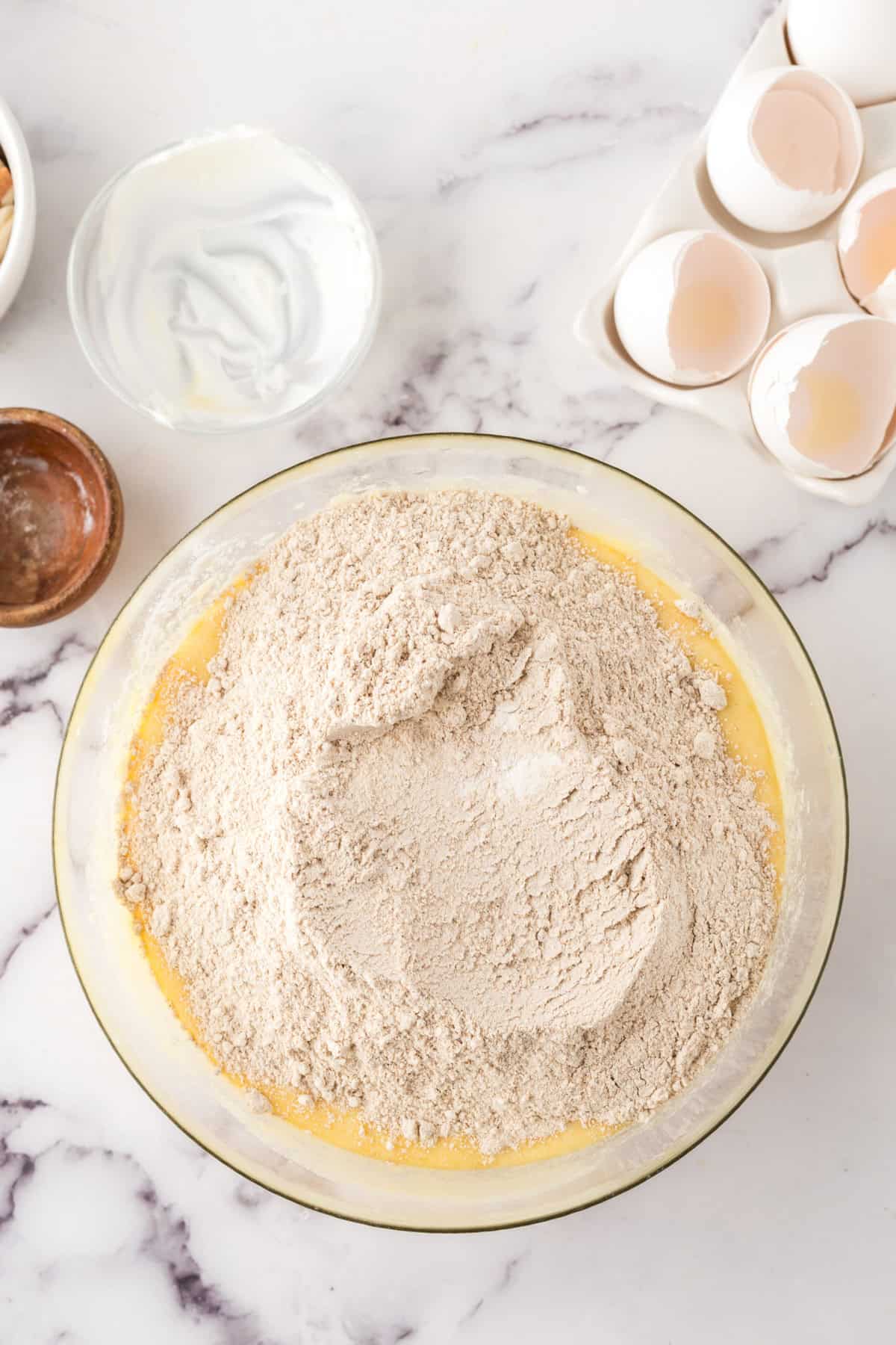A glass mixing bowl with flour atop wet yellow batter, surrounded by cracked eggshells, a small bowl of yogurt, and a wooden bowl on a white marble countertop—perfect for starting your favorite honey cake recipe.