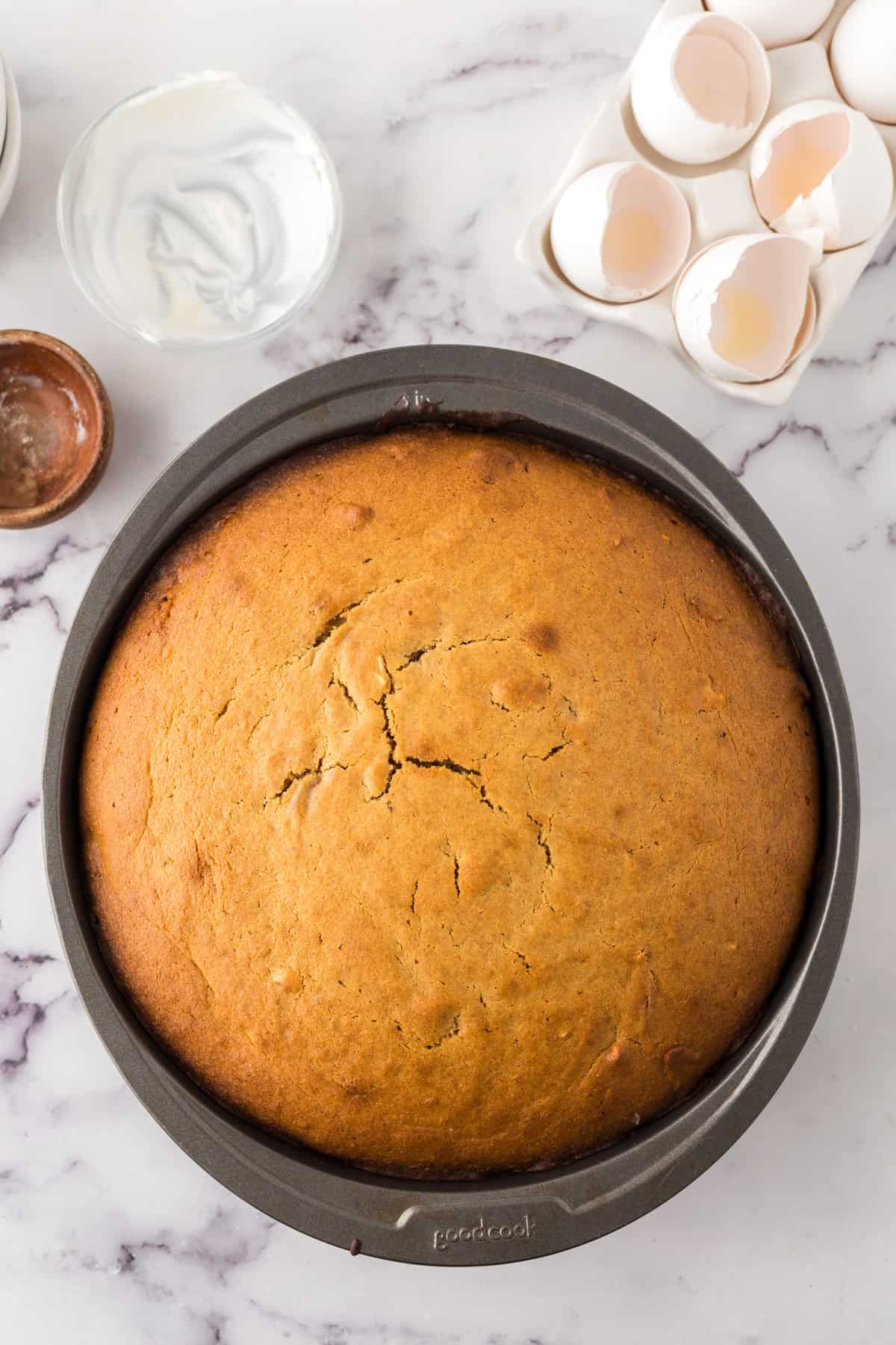 A round, golden-brown honey cake rests in a baking pan on a marble counter, surrounded by a bowl of yogurt, a small dish, and a carton with cracked eggshells—ingredients for the perfect honey cake recipe.