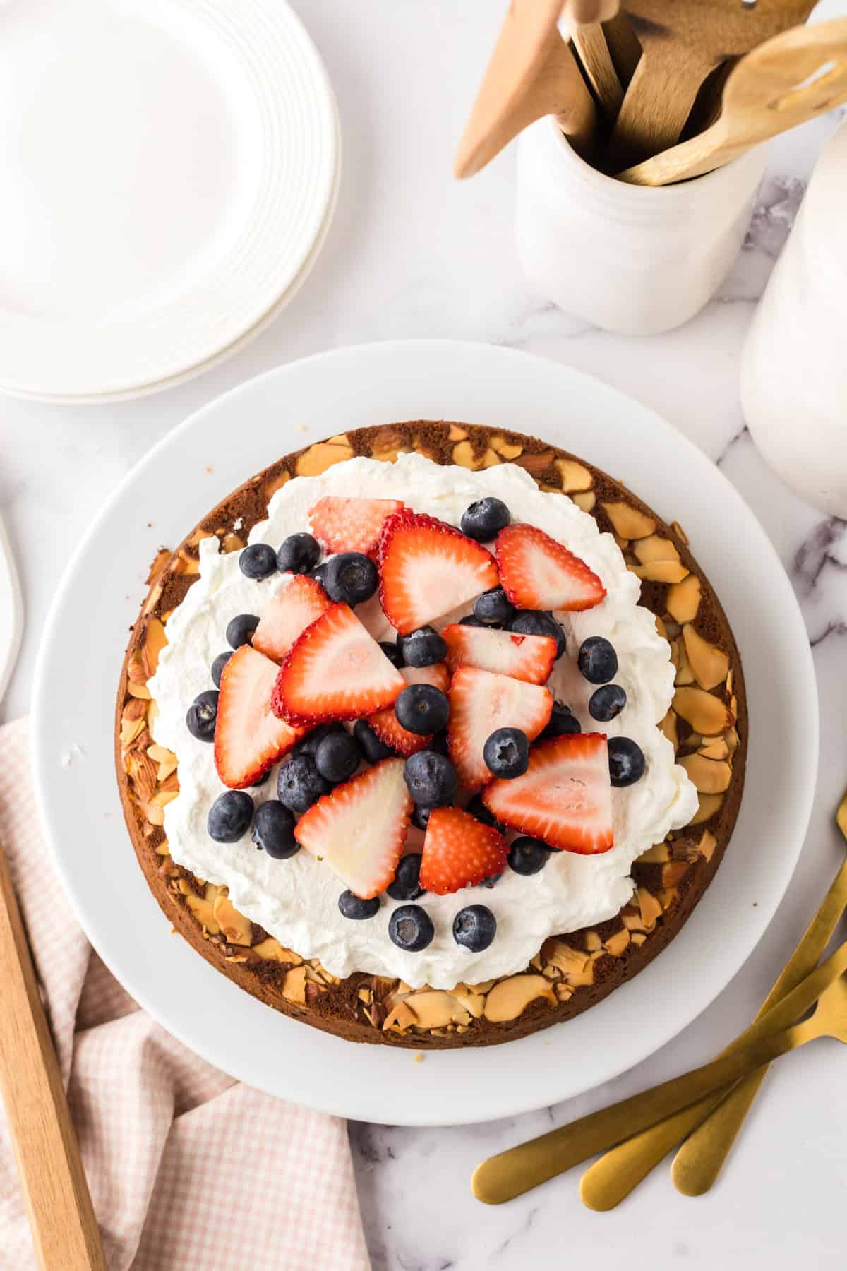 A round honey cake recipe comes to life, topped with whipped cream, sliced strawberries, and blueberries on a white plate. The cake’s edges are decorated with sliced almonds, set beside gold utensils, a napkin, and wooden kitchen tools.