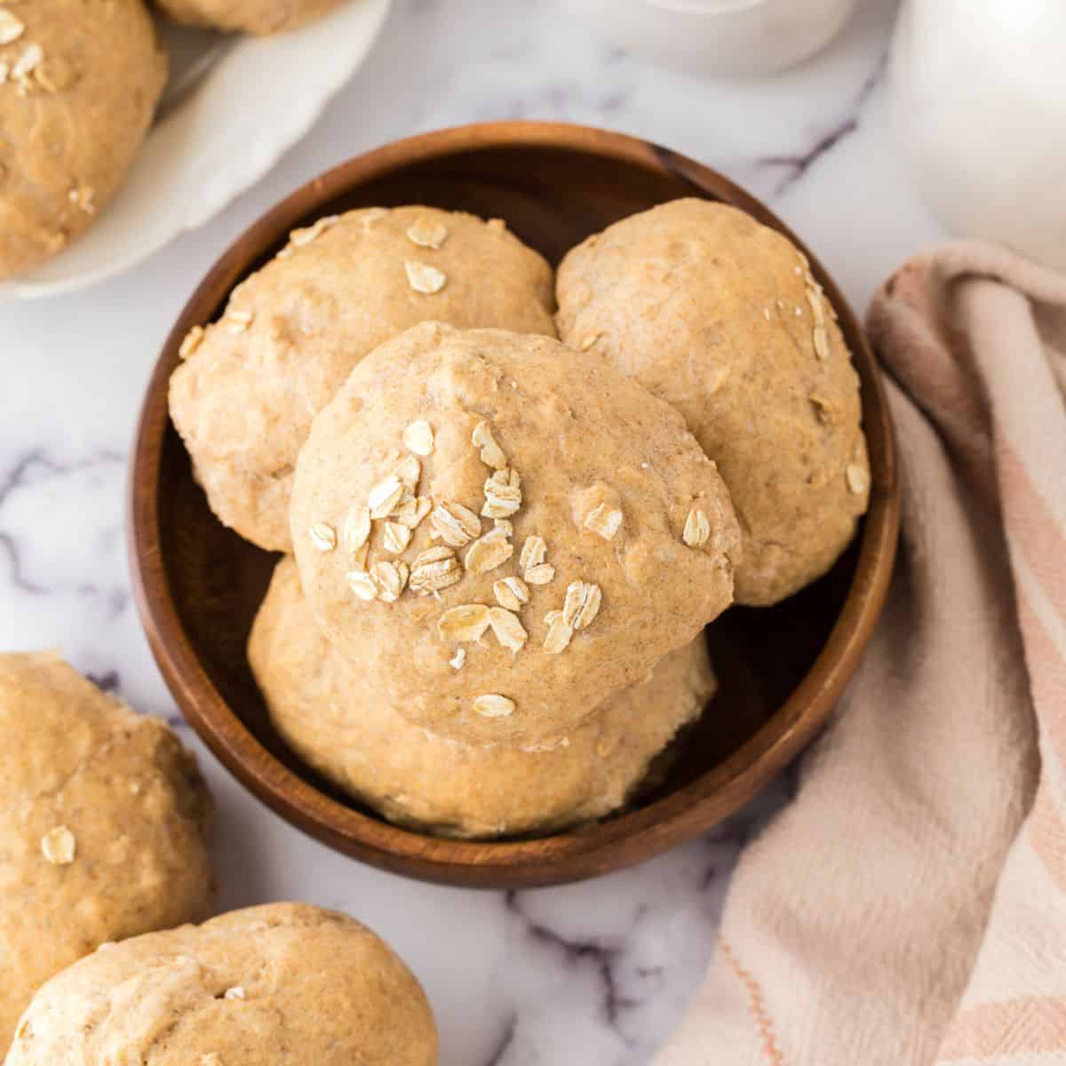 A wooden bowl filled with round, homemade whole wheat hamburger buns topped with oats. The bowl rests on a marble surface, accompanied by a beige cloth and additional rolls nearby, creating a rustic and cozy presentation.