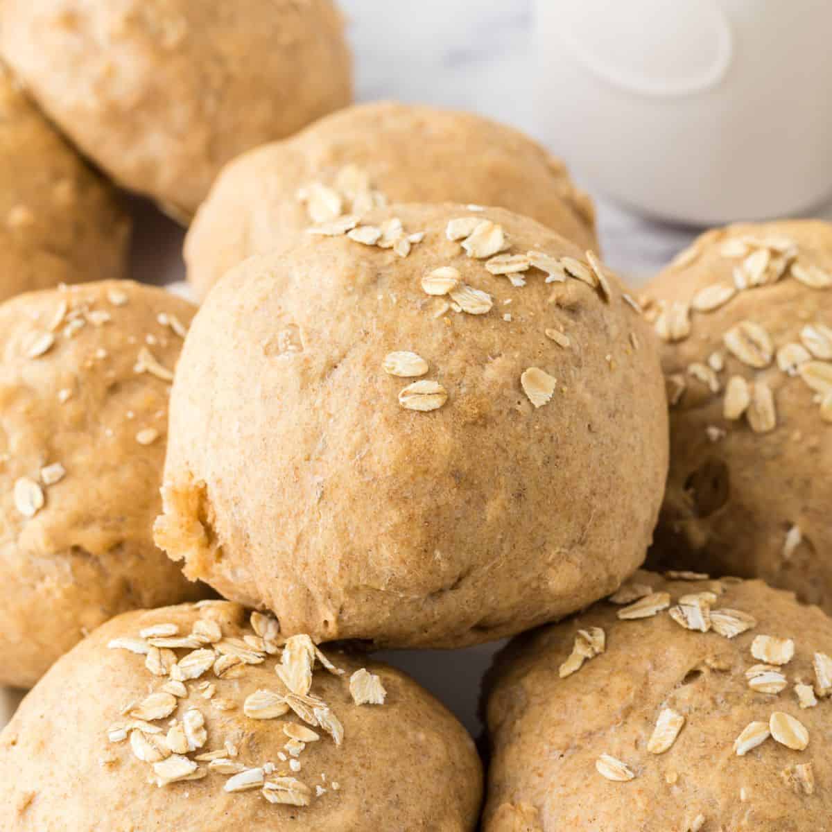 A close-up of freshly baked whole wheat hamburger buns topped with oats. The buns are stacked on a marble surface with a hint of a white, round container in the background.