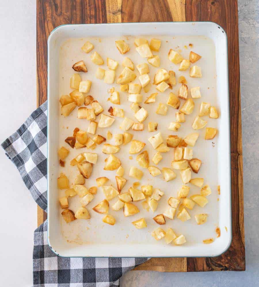 Crispy oven roasted potatoes on a white enamel baking sheet resting on a wooden board.