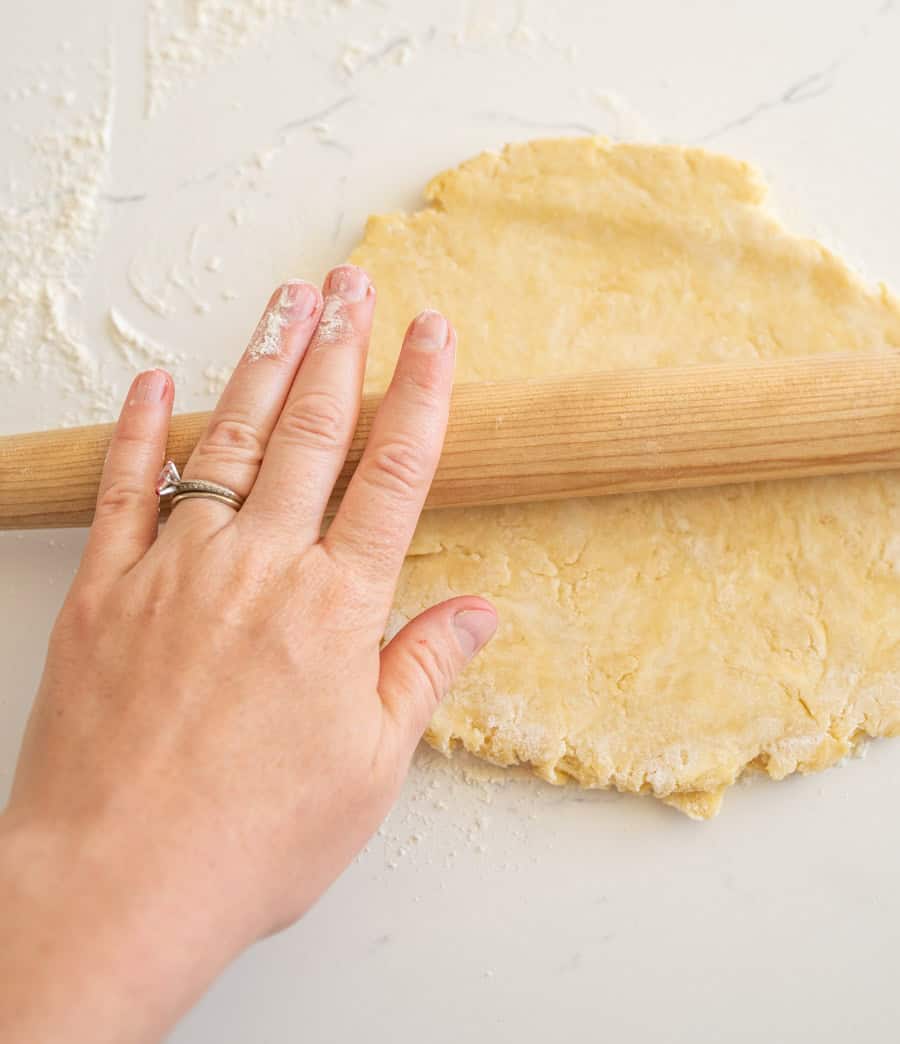 A hand using a rolling pin to roll out a pie crust on a lightly floured white counter.