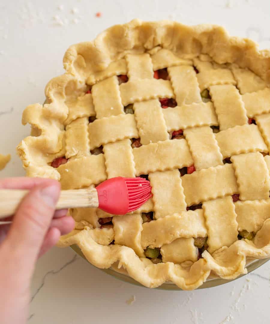 A hand holding a red silicone brush brushing water onto the lattice pie crust top.