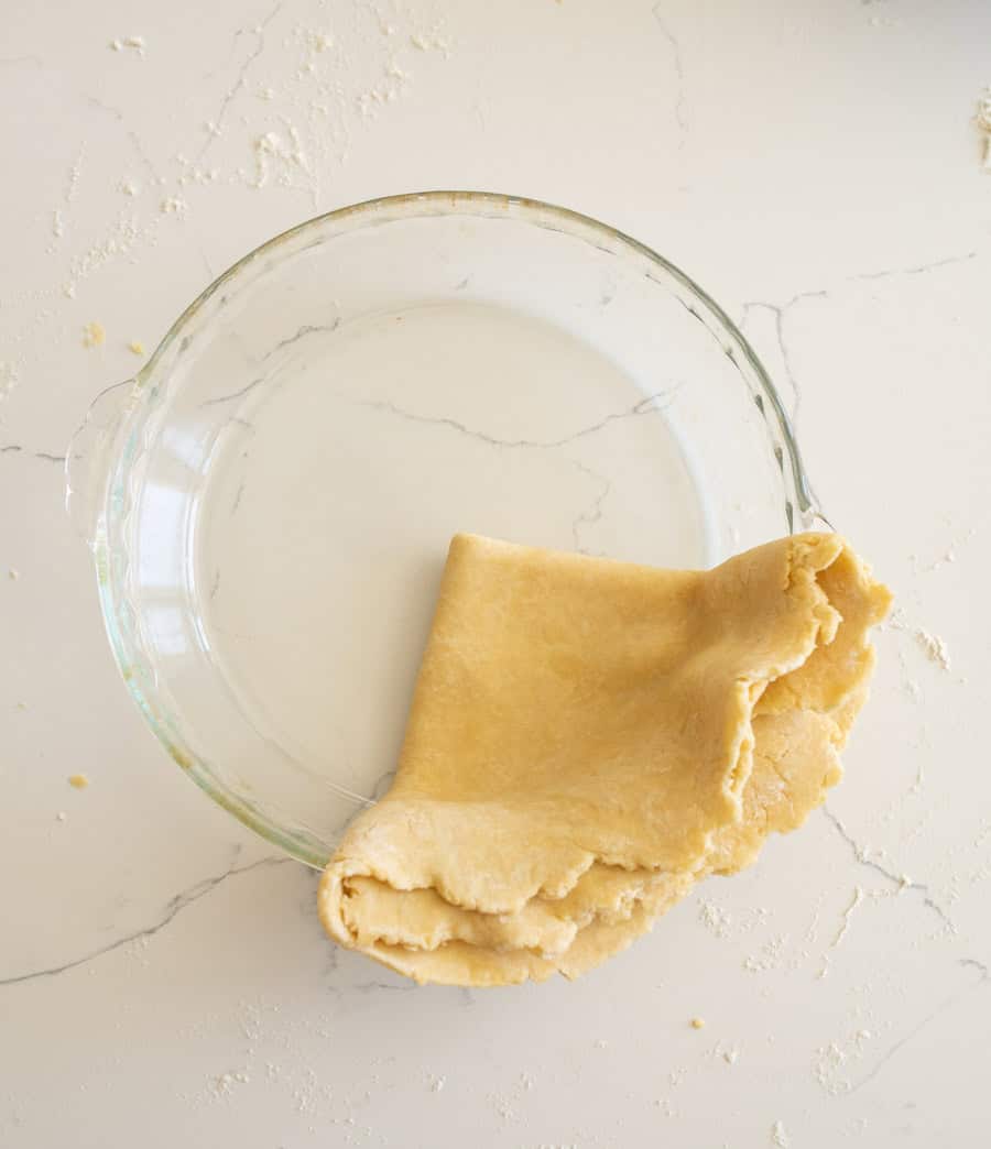 The pie crust folded twice into a quarter of a circle in a clear glass pie plate on a white counter.
