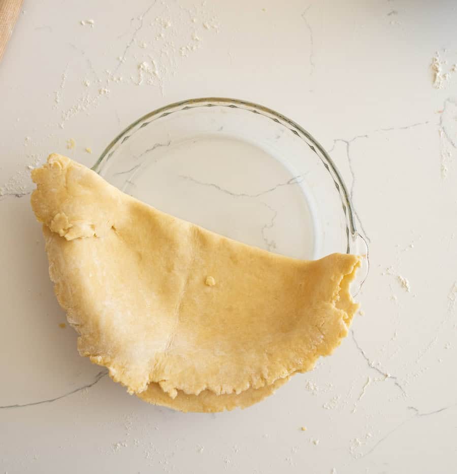 The pie crust unfolded into a semi-circle in a clear glass pie plate on a white counter.