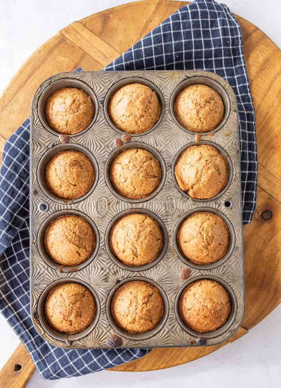 A dozen baked sourdough muffins on a blue and white towel on wooden cutting board.