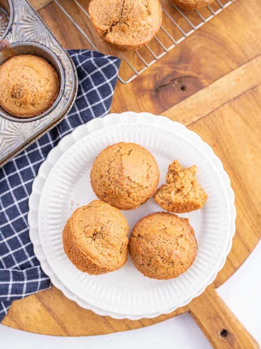 Three sourdough muffins on a white plate with baked muffins in a muffin pan and baked muffins on a cooling rack in the background.