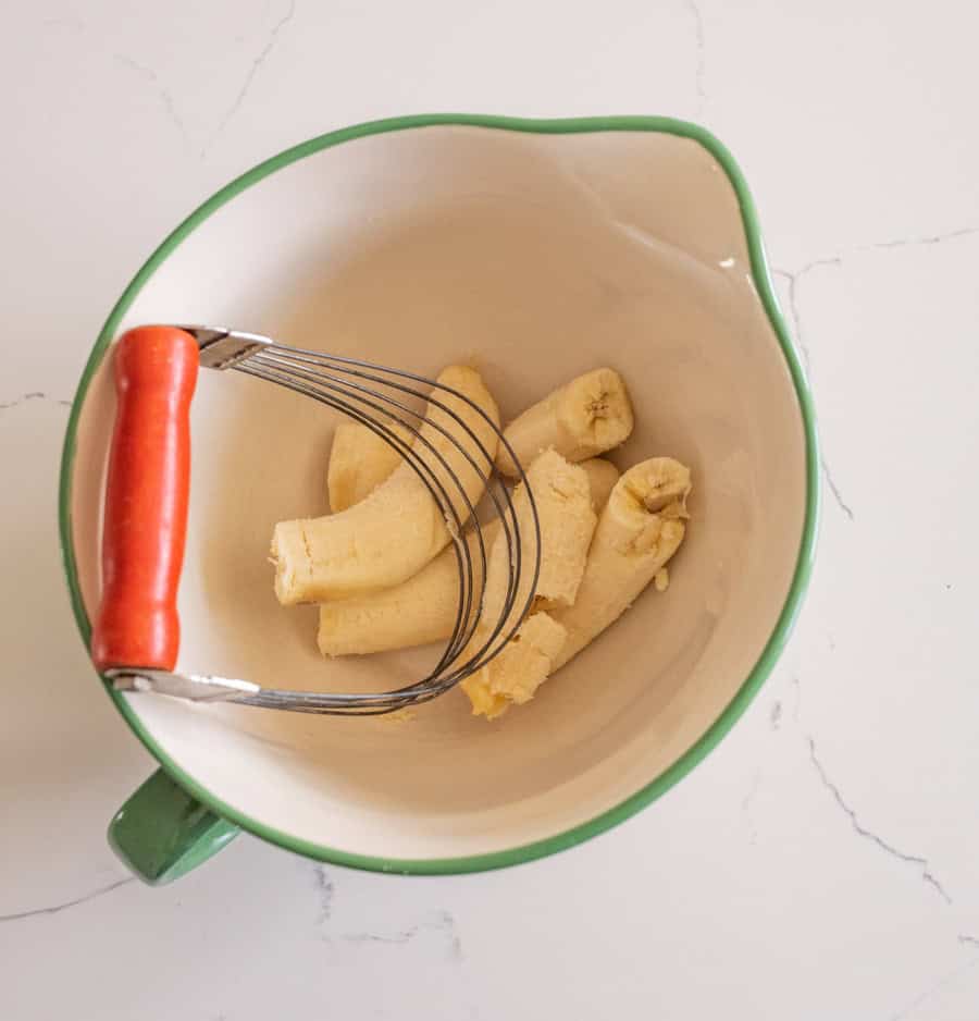 Three bananas in a white bowl with a green rim and a pastry blender with a red handle on a white counter.