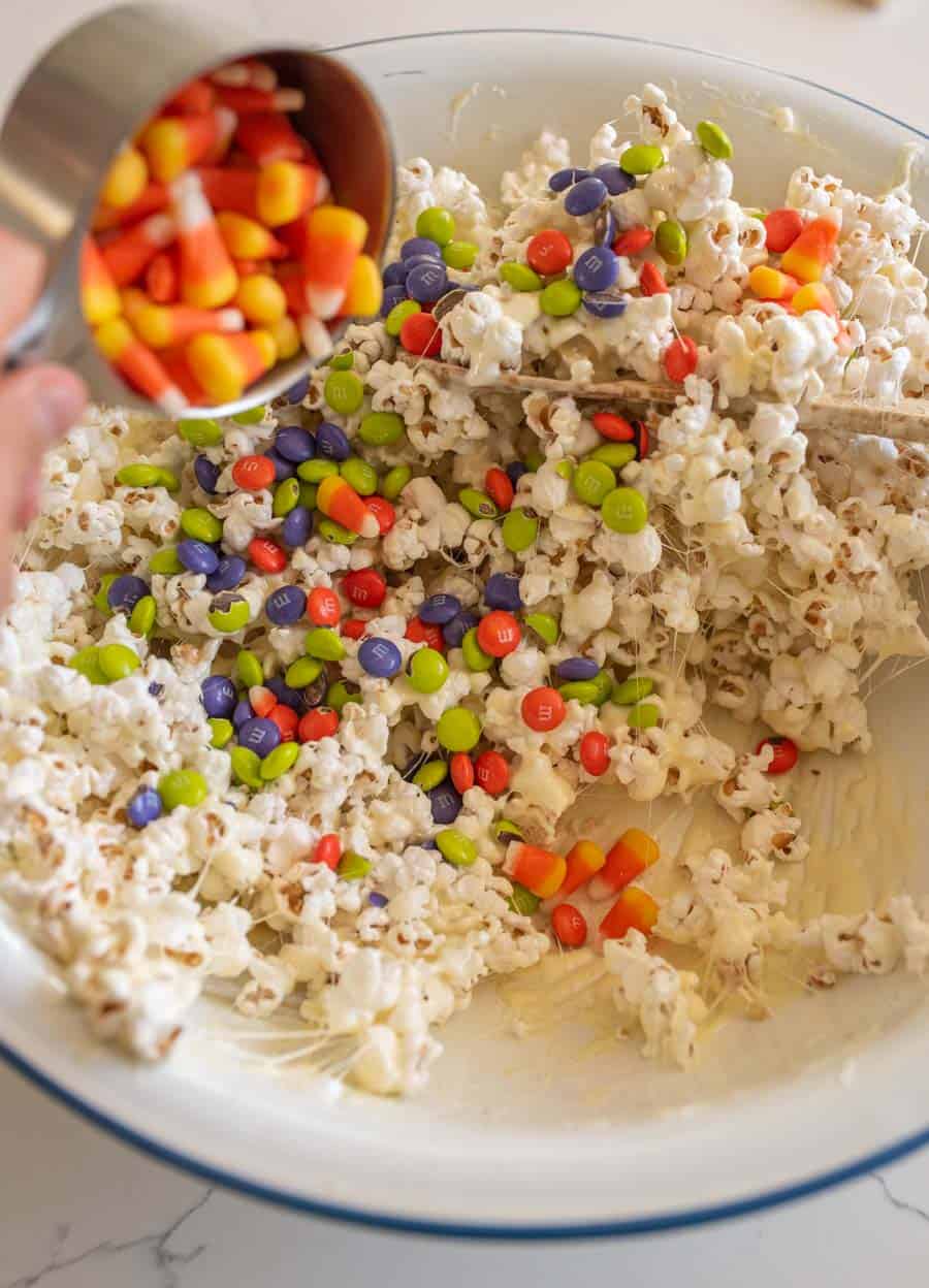 White bowl with popcorn coated in the melted marshmallow mixture with M&Ms and a small measuring cup pouring in the candy corn.