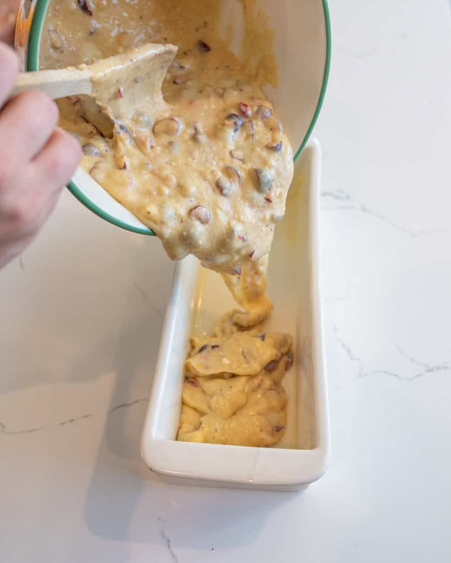 Cranberry orange bread batter being poured into a white ceramic tea cake pan using a wooden spoon.