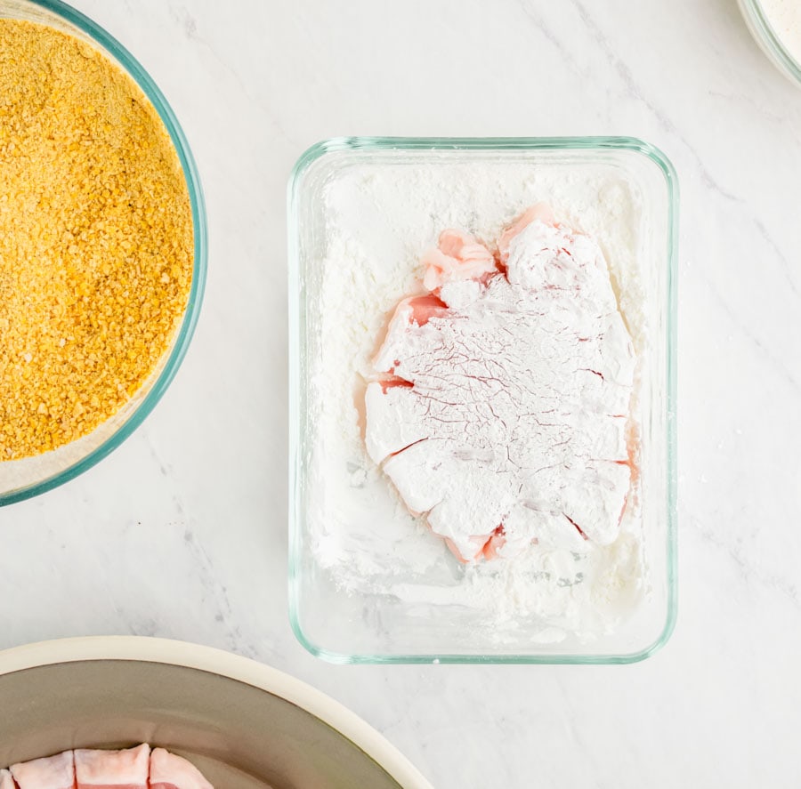 Pork chop being dredged in the corn starch in a shallow clear glass container.