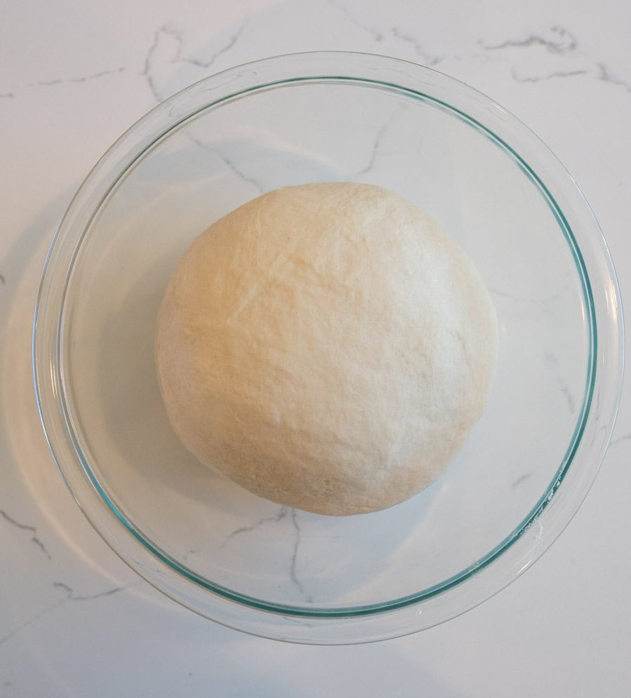 Ball of dough in a clear glass bowl on a white marble counter.