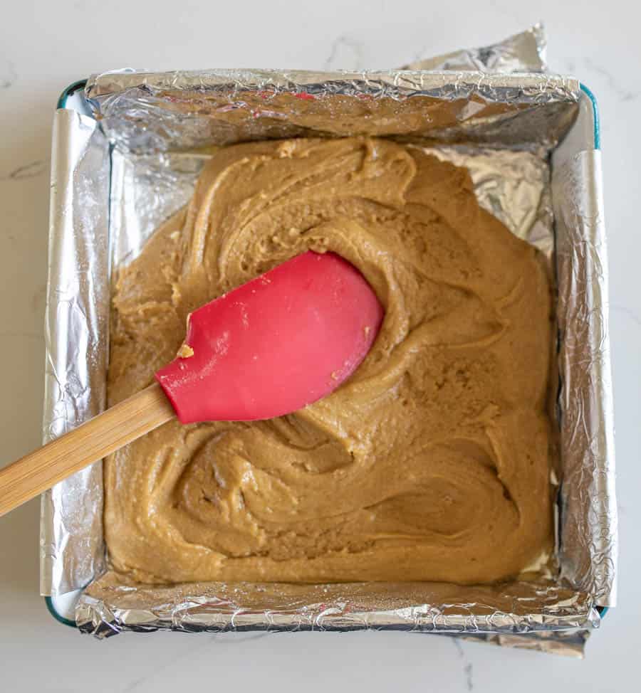 Halloween candy cookie bar batter being spread with a red spatula with a wooden handle in a 9 by 9 inch pan.