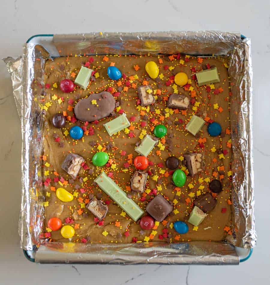 A pan of Halloween candy cookie bar batter with chopped up pieces of Halloween candy on top.