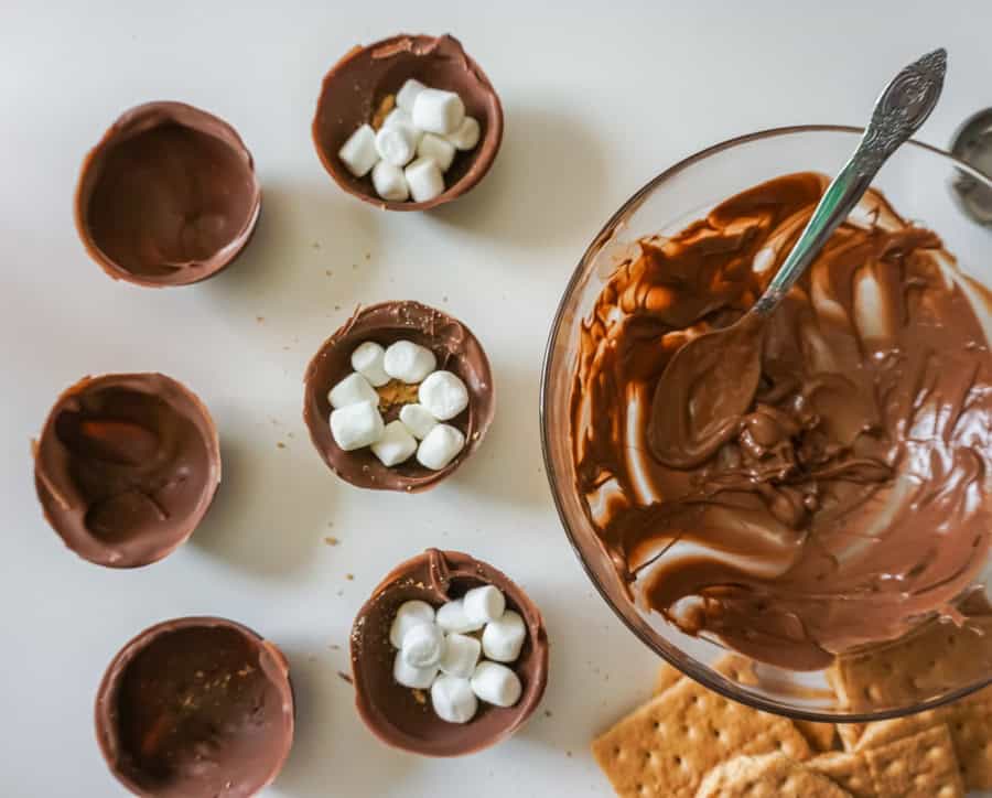 Half of a hot chocolate bomb filled with hot cocoa and mini marshmallows next to empty halves and a clear glass bowl with melted chocolate.