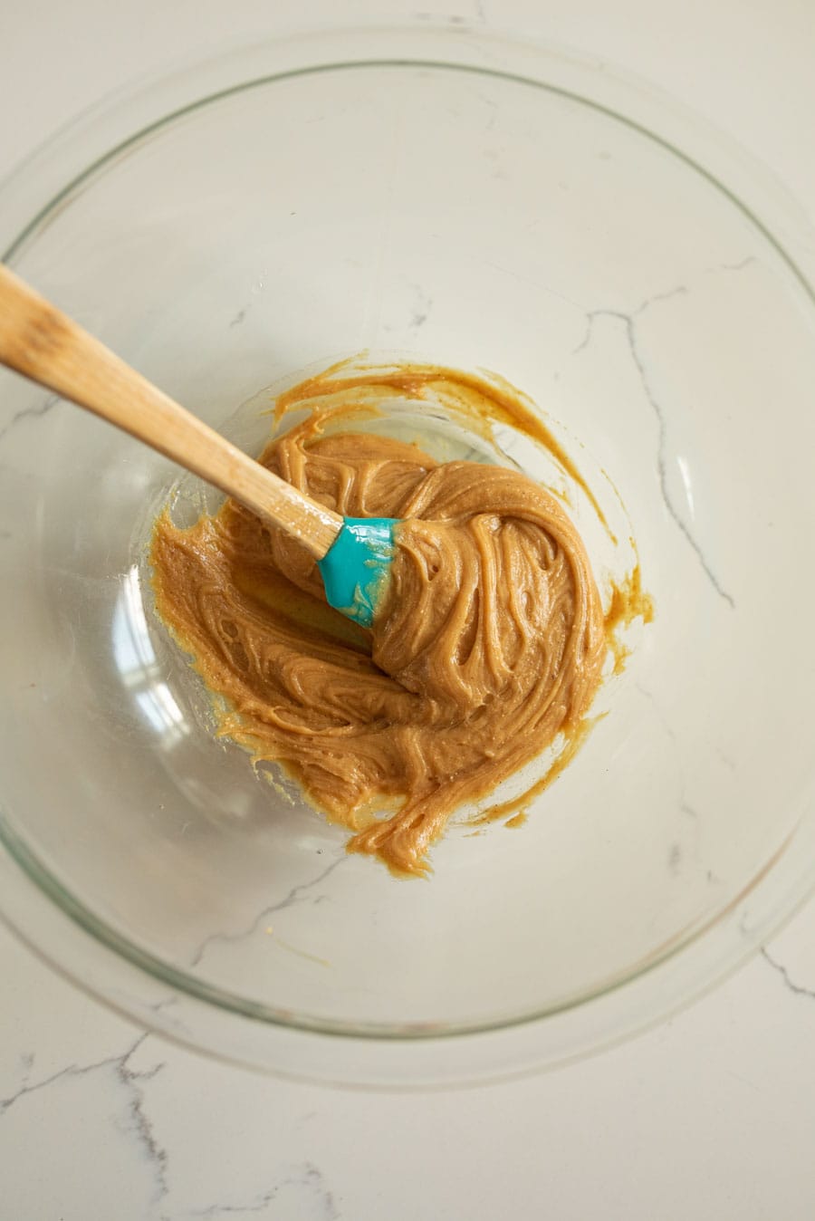 A clear glass mixing bowl with honey and peanut butter being mixed together with a spatula with a wooden handle.