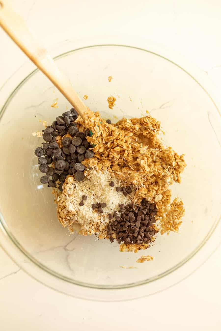All of the ingredients for one of the no-bake oatmeal energy ball recipes in a clear glass bowl with a spatula with a wooden handle.