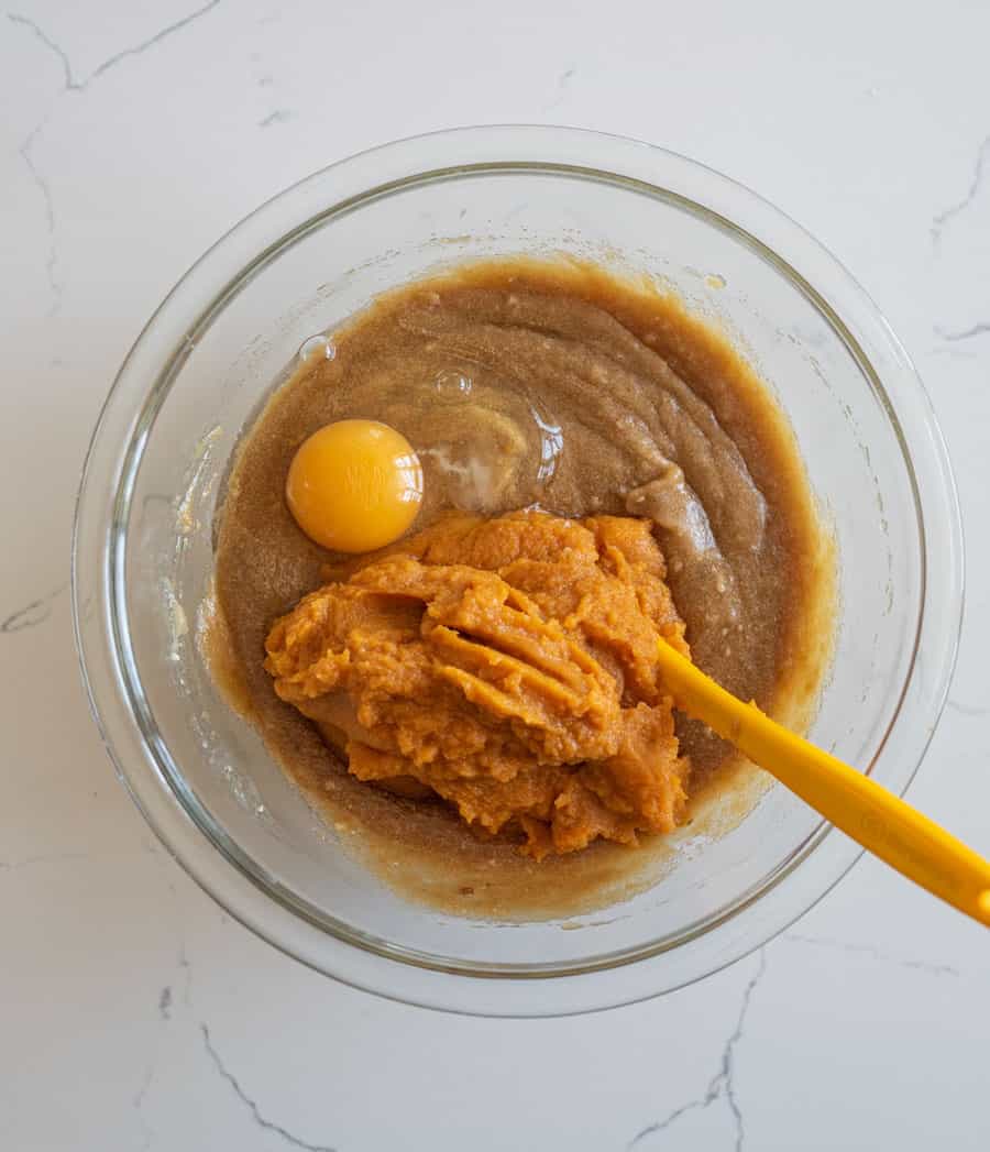 An egg and pumpkin added to the pumpkin cake batter in a clear glass bowl on a white counter.