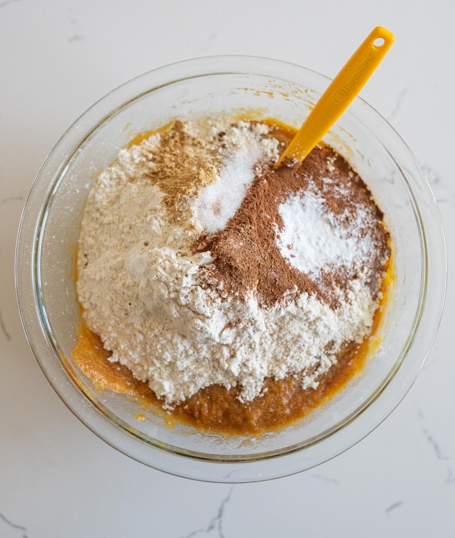 Dry ingredients added to the pumpkin cake batter in a clear glass bowl on a white counter.