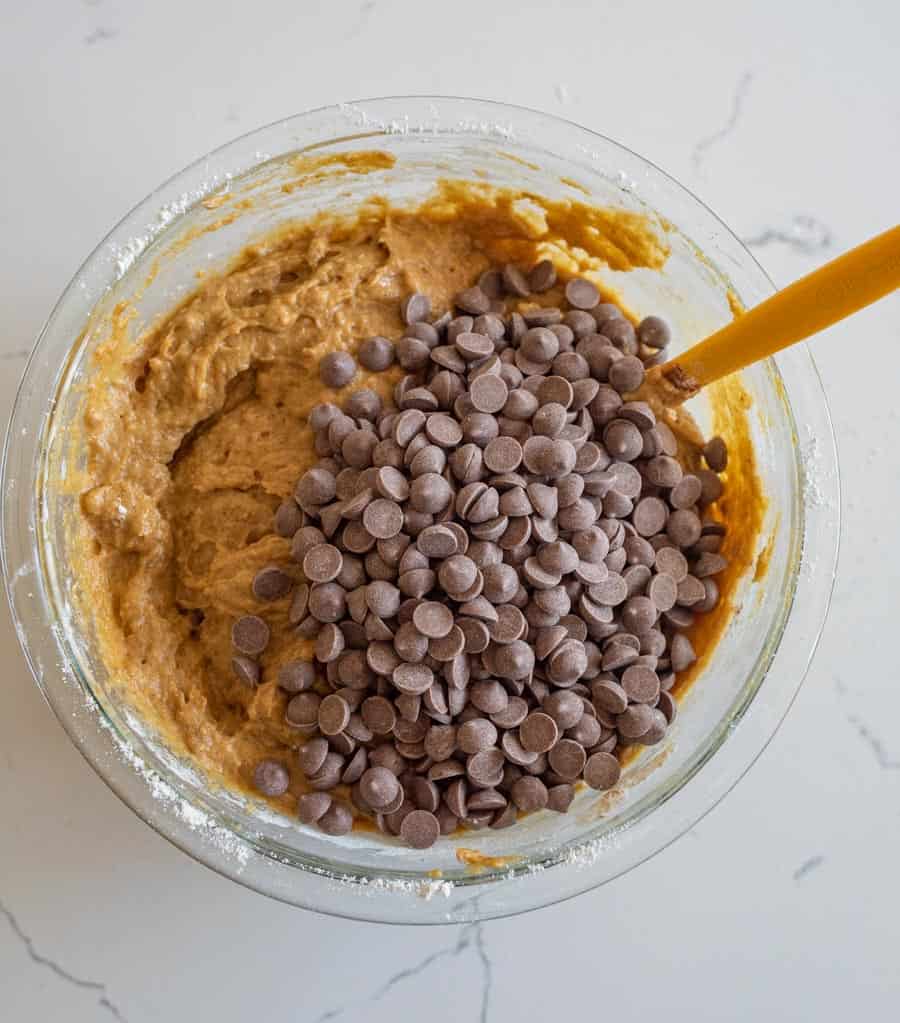 Chocolate chips added to the pumpkin cake batter in a clear glass bowl on a white counter.