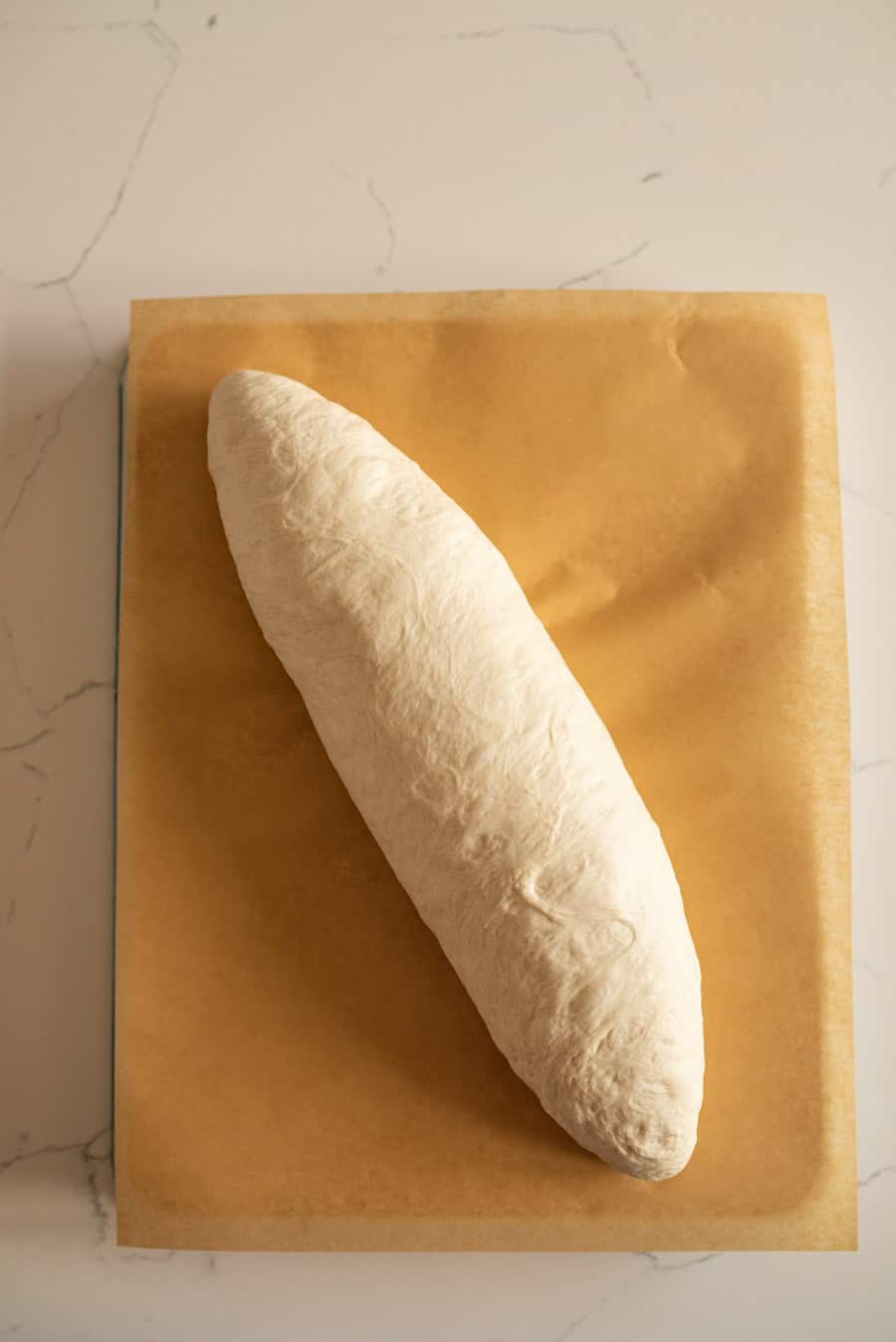 Italian bread dough on brown parchment paper after being shaped into a loaf.