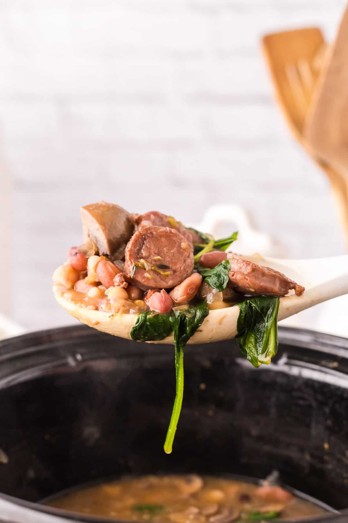 crockpot with sausage bean soup with spinach with being scooped with a white spoon.