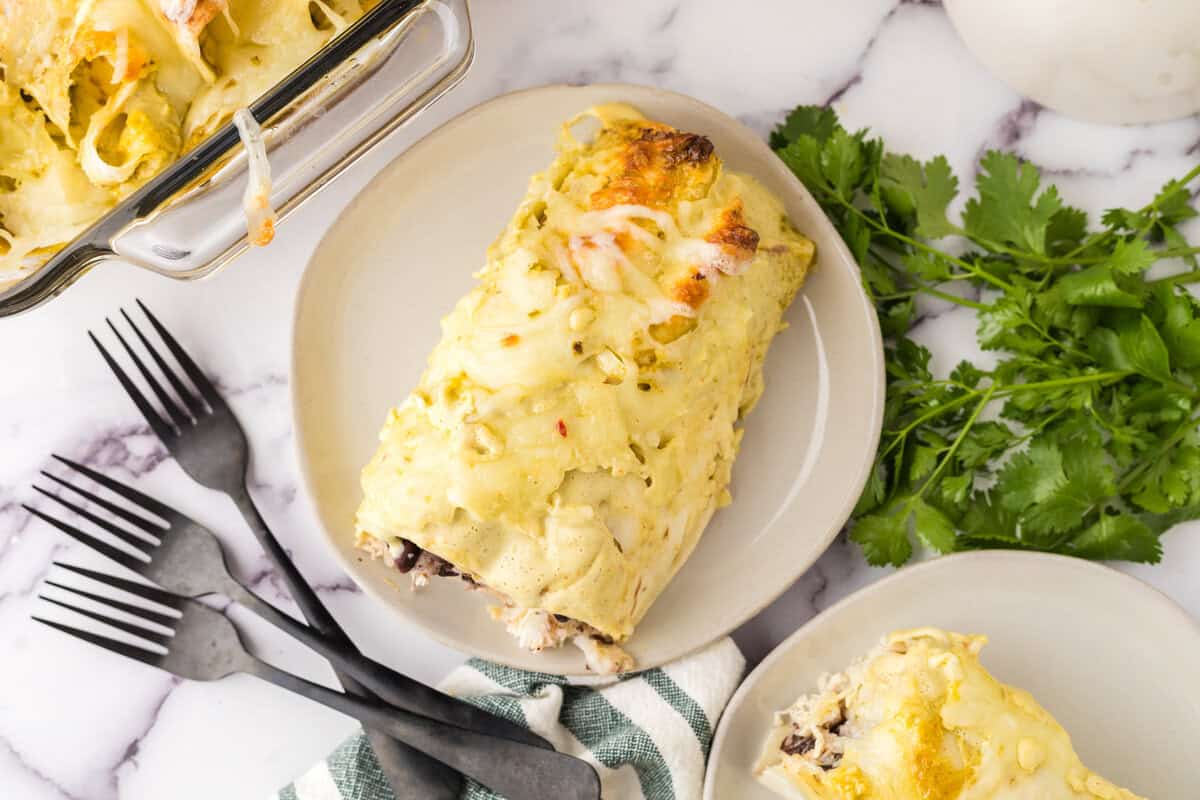 A plate with a cheesy enchilada covered in green sauce sits next to a casserole dish, a bunch of fresh cilantro, and three black forks on a marble surface.