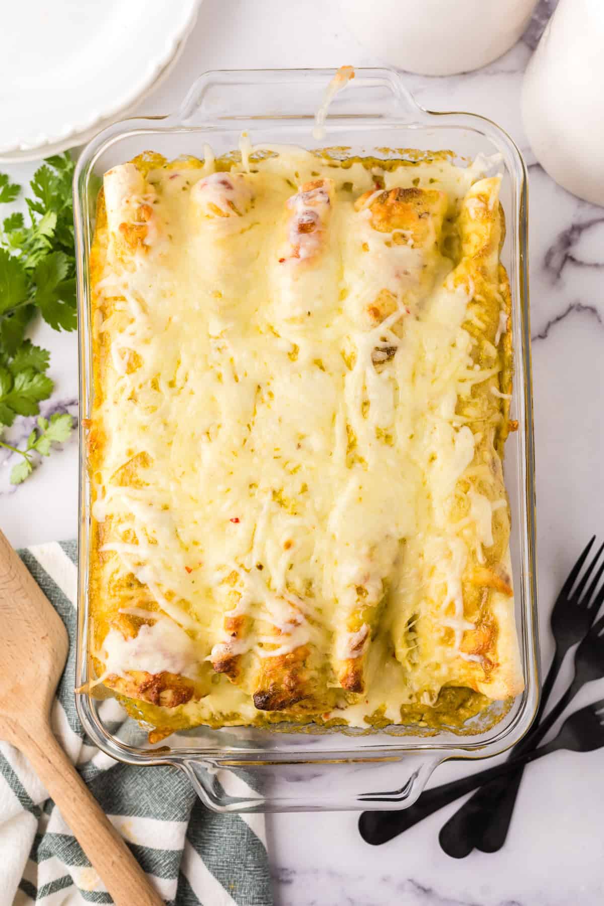 A glass baking dish filled with baked enchiladas, reminiscent of a chicken burrito recipe, topped with melted cheese on a marble surface next to black forks, a wooden spoon, a striped towel, and fresh cilantro.