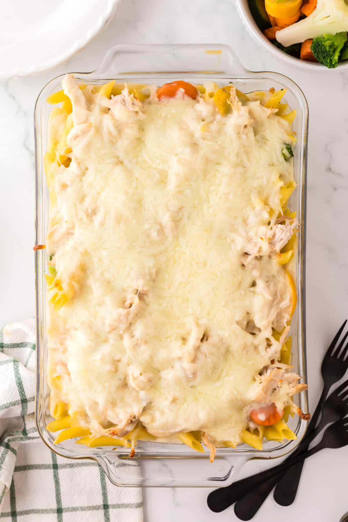 A glass baking dish filled with a cheesy chicken vegetable pasta casserole, topped with melted cheese and visible noodles and veggies. A striped towel, utensils, and a bowl of mixed vegetables are nearby on a white surface.