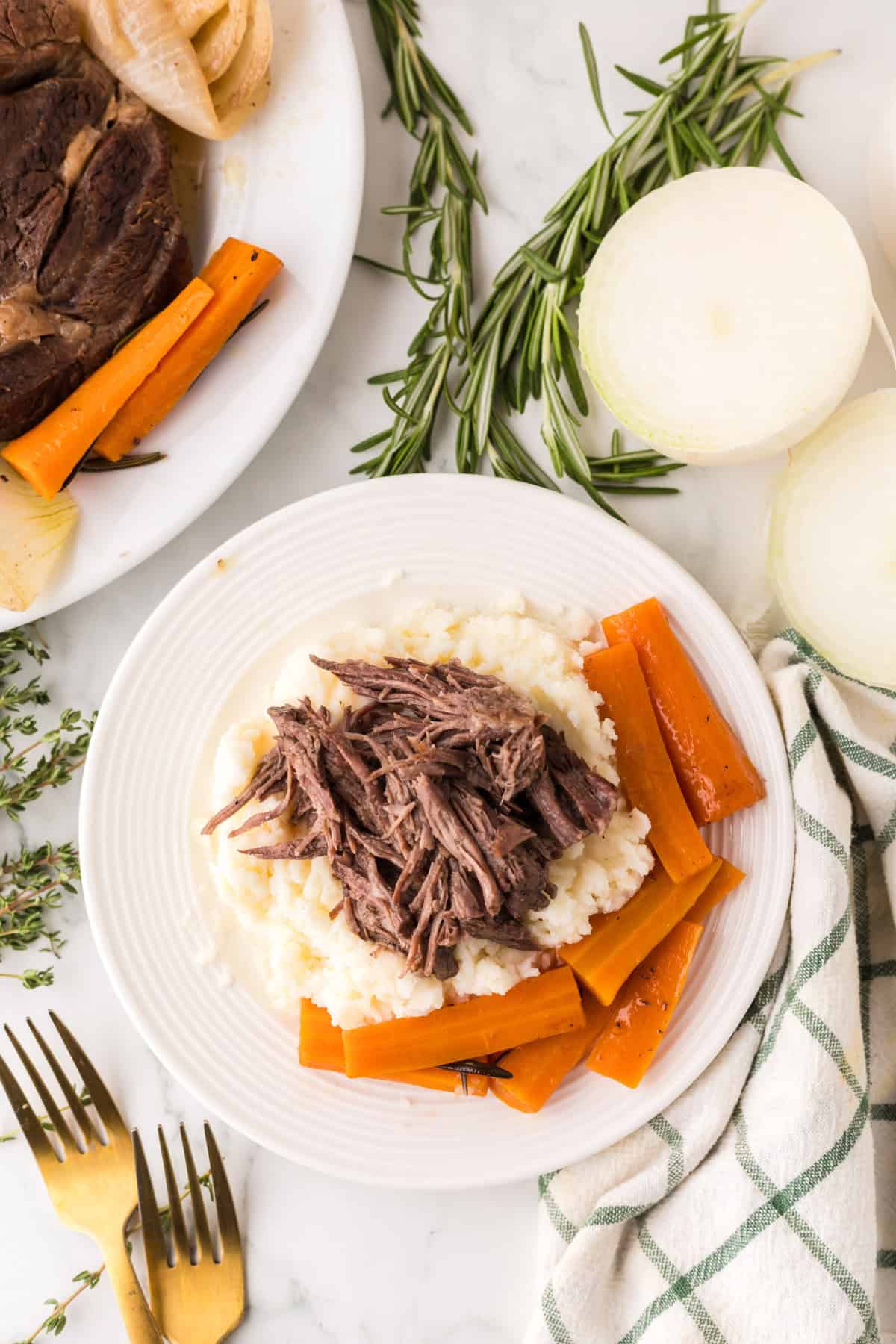 A plate of mashed potatoes topped with tender shredded pot roast—perfect if you make pot roast in a slow cooker—is surrounded by cooked carrot sticks, herbs, and onions on a white surface. A second plate with more roast sits nearby.