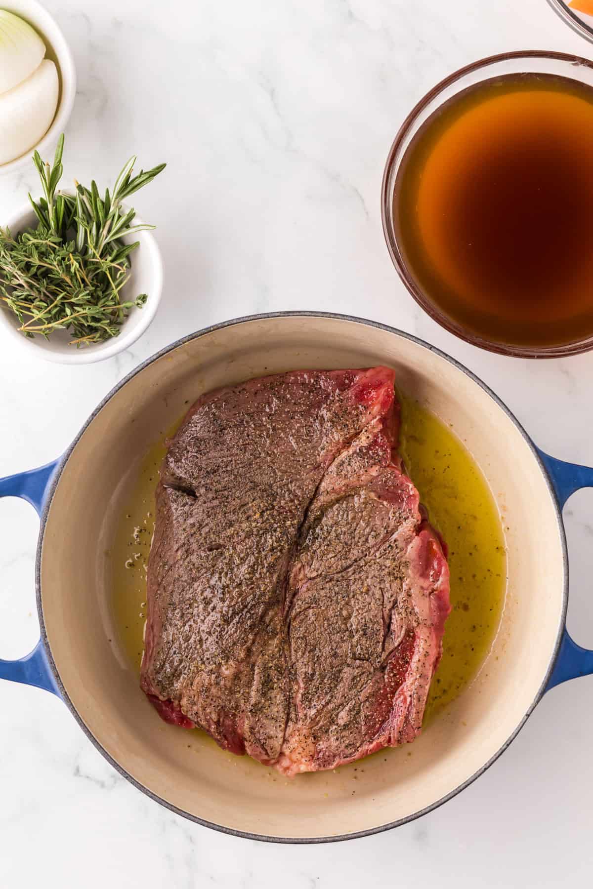 Raw, seasoned beef roast sits in a blue Dutch oven with oil. Nearby are a bowl of brown broth, fresh herbs, and part of a sliced onion—perfect ingredients if you want to make pot roast in slow cooker style at home.