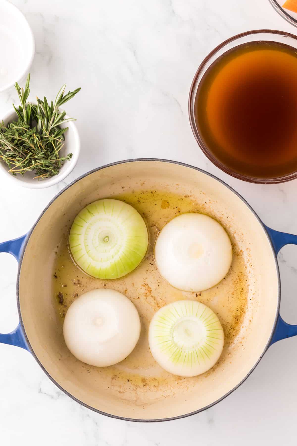 A blue pot with four halved onions sautéing in oil, next to a brown liquid in a bowl and a small dish of fresh rosemary—perfect ingredients if you want to make pot roast in slow cooker style on a white marble surface.