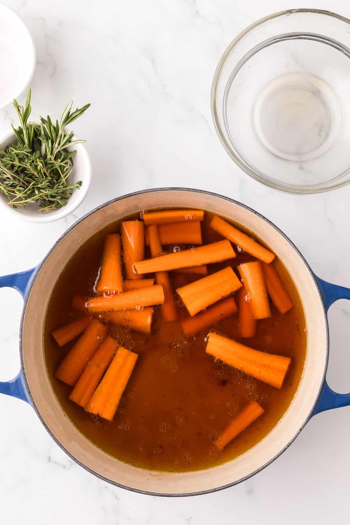 A blue pot filled with carrot sticks cooking in broth sits on a white surface. Nearby are a small bowl of fresh rosemary and an empty glass bowl, perfect for those who want to make pot roast in slow cooker style at home.