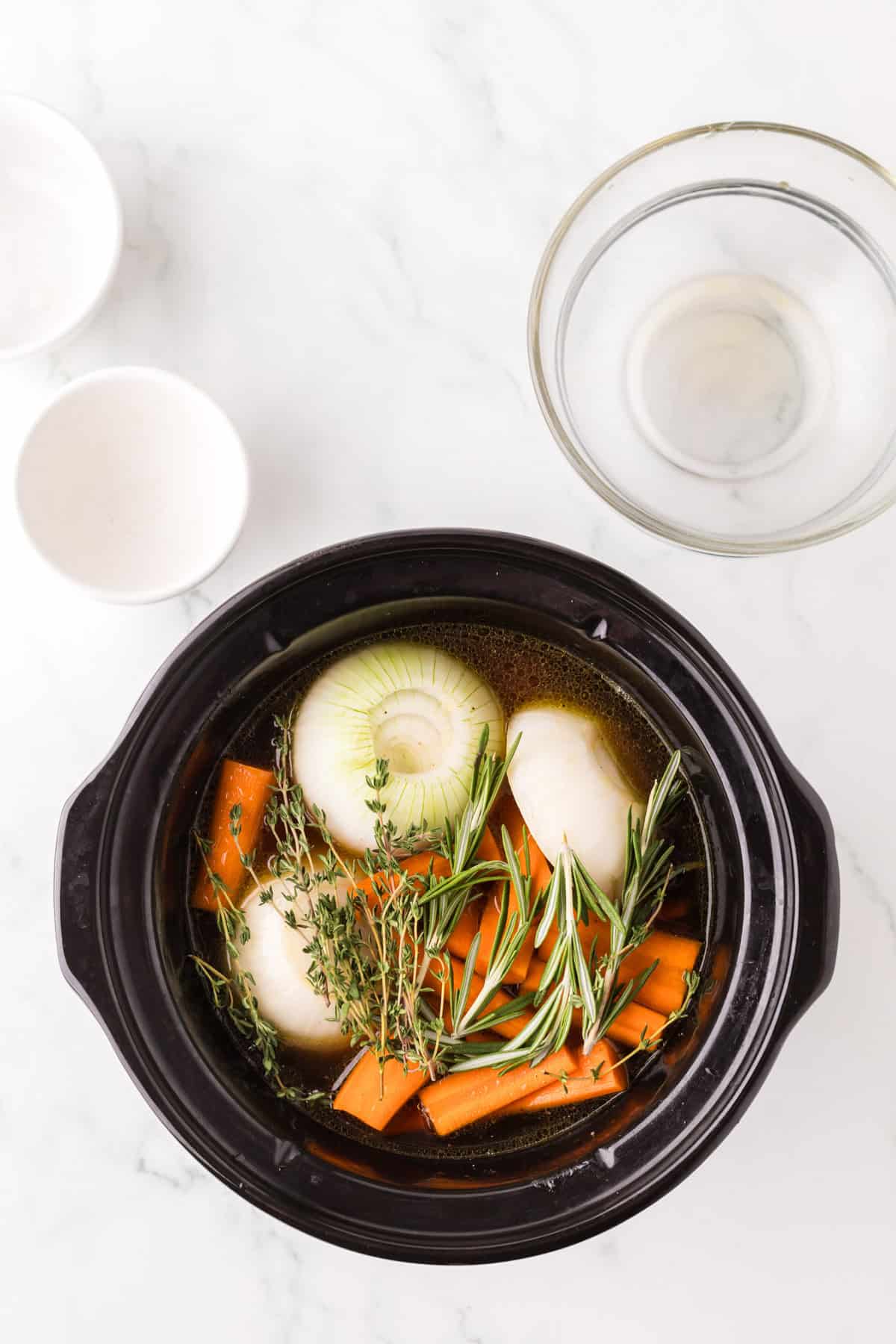 A slow cooker filled with halved onions, chopped carrots, fresh thyme, and rosemary in broth sits on a white countertop—perfect ingredients to make pot roast in slow cooker—near empty bowls and a clear glass bowl.