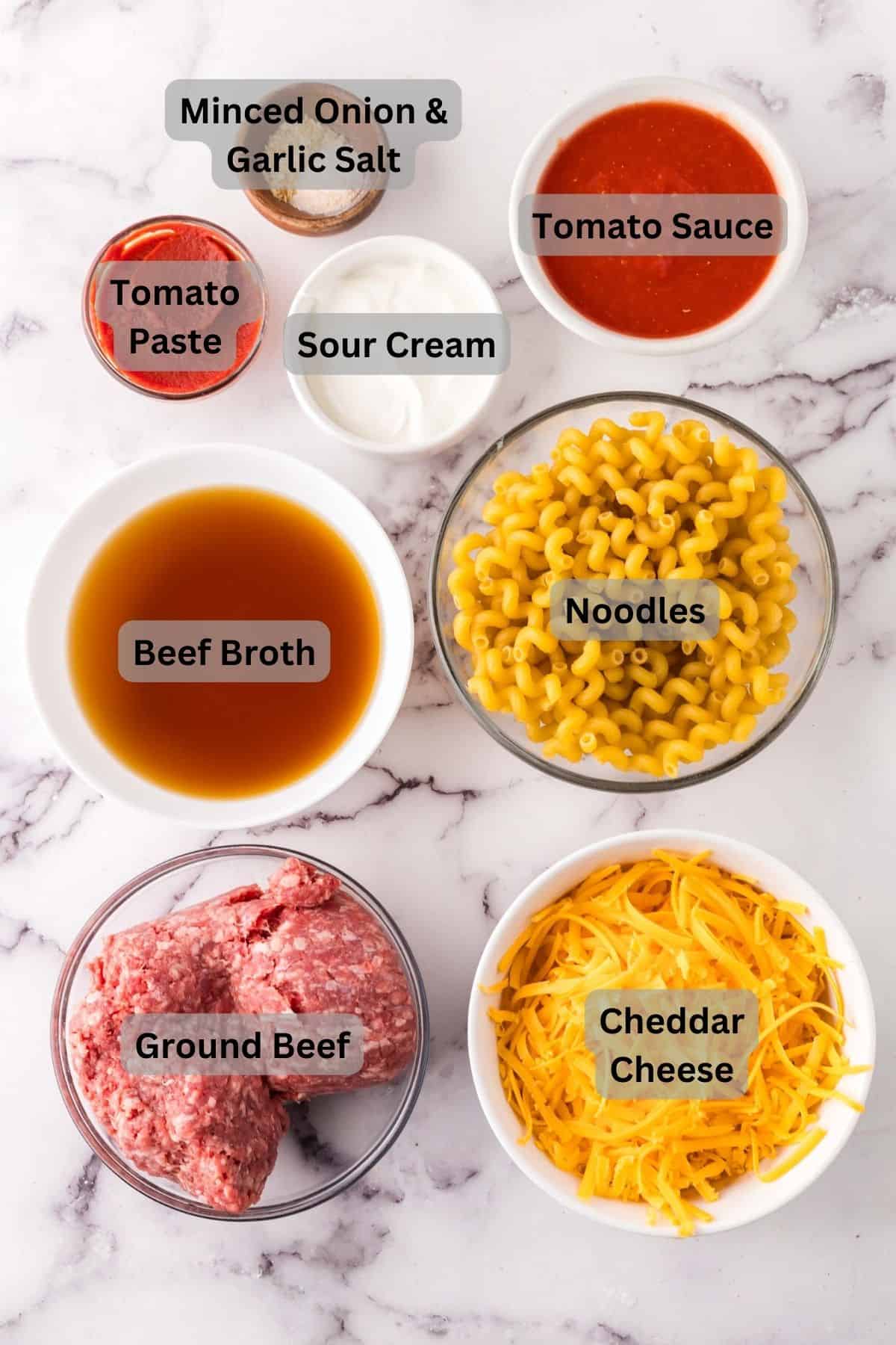 Top view of bowls with ingredients for cheeseburger casserole: minced onion and garlic salt, tomato paste, sour cream, tomato sauce, beef broth, uncooked macaroni noodles, ground beef, and shredded cheddar cheese on a white surface.