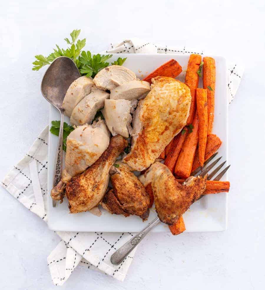 A white plate with roasted chicken pieces and seasoned carrot sticks, garnished with parsley. Perfect for an easy weekly meal plan. A serving spoon and fork rest on the plate, which sits on a white checkered napkin.