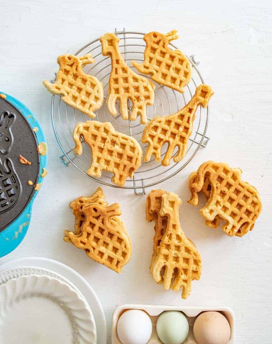 Animal-shaped waffles, including giraffes, elephants, and rhinos, are arranged on a cooling rack and white surface—perfect for adding fun to any easy weekly meal plan. Nearby sit a waffle maker, empty plates, and a carton with three eggs.