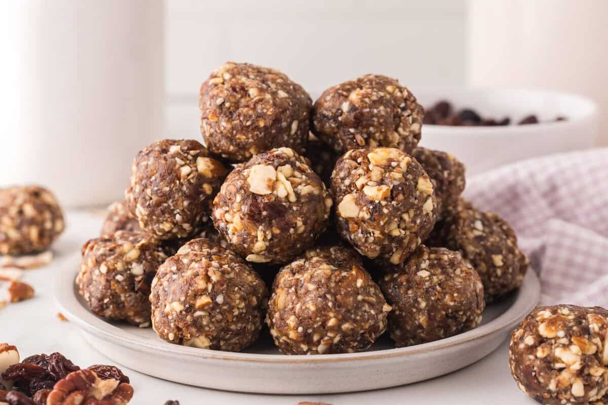 A plate stacked with round, brown energy bites made from chopped nuts and dried fruit—perfect for an easy weekly meal plan—with more bites and ingredients scattered around on a white surface.