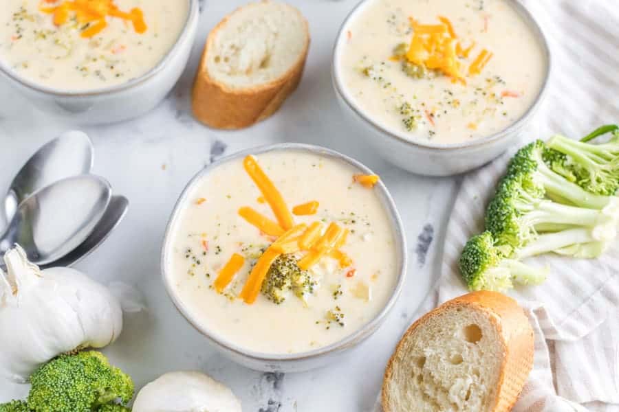 Three bowls of creamy broccoli cheddar soup topped with shredded cheese, surrounded by sliced baguette, garlic, and broccoli florets—an ideal pick for any easy weekly meal plan. Two spoons rest on the white marble surface.