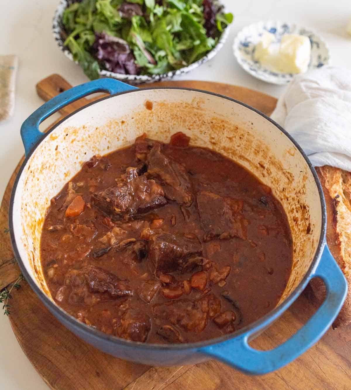 A blue Dutch oven filled with julia child’s beef bourguignon sits on a wooden board, with a bowl of mixed green salad and a plate of butter in the background. Slices of bread are visible beside the pot.