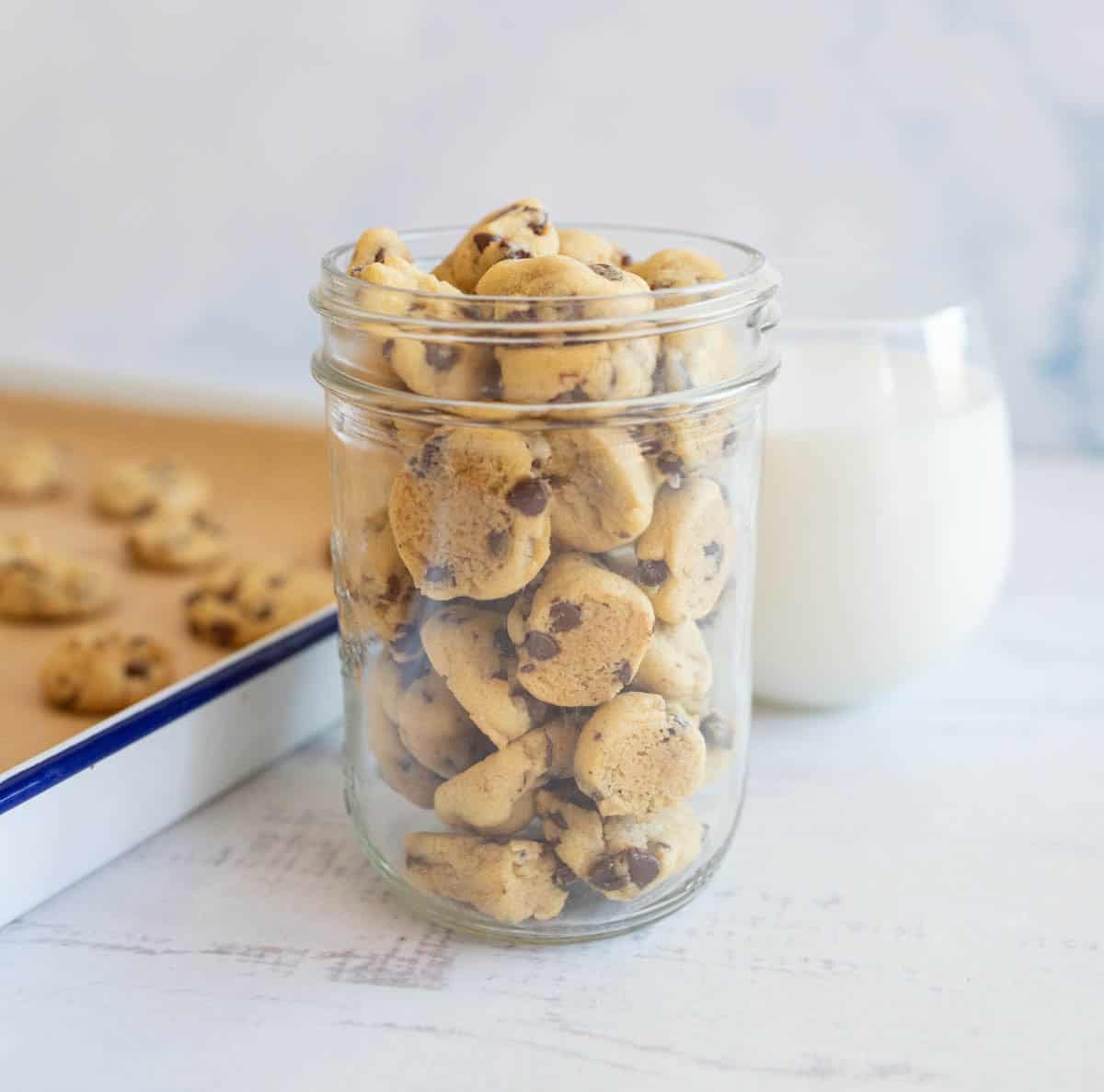 A jar brimming with delicious mini chocolate chip cookies rests on a white surface. Behind it, a glass of milk and a baking tray piled high with more cookies complete the delightful scene, set against a soft, light background.