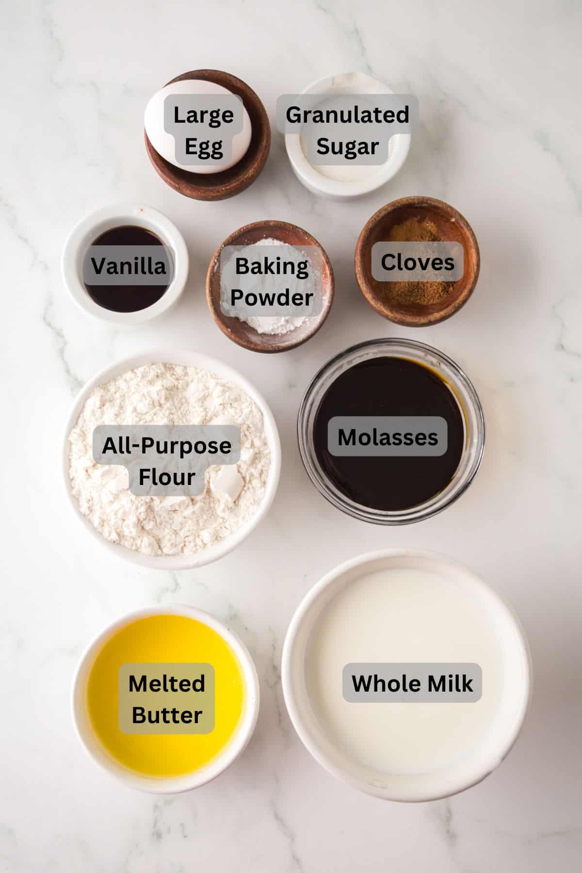 Top-down view of nine ingredients in separate bowls on a white surface. Ingredients include a large egg, granulated sugar, vanilla, baking powder, cloves, all-purpose flour, molasses, melted butter, and whole milk, each labeled accordingly.