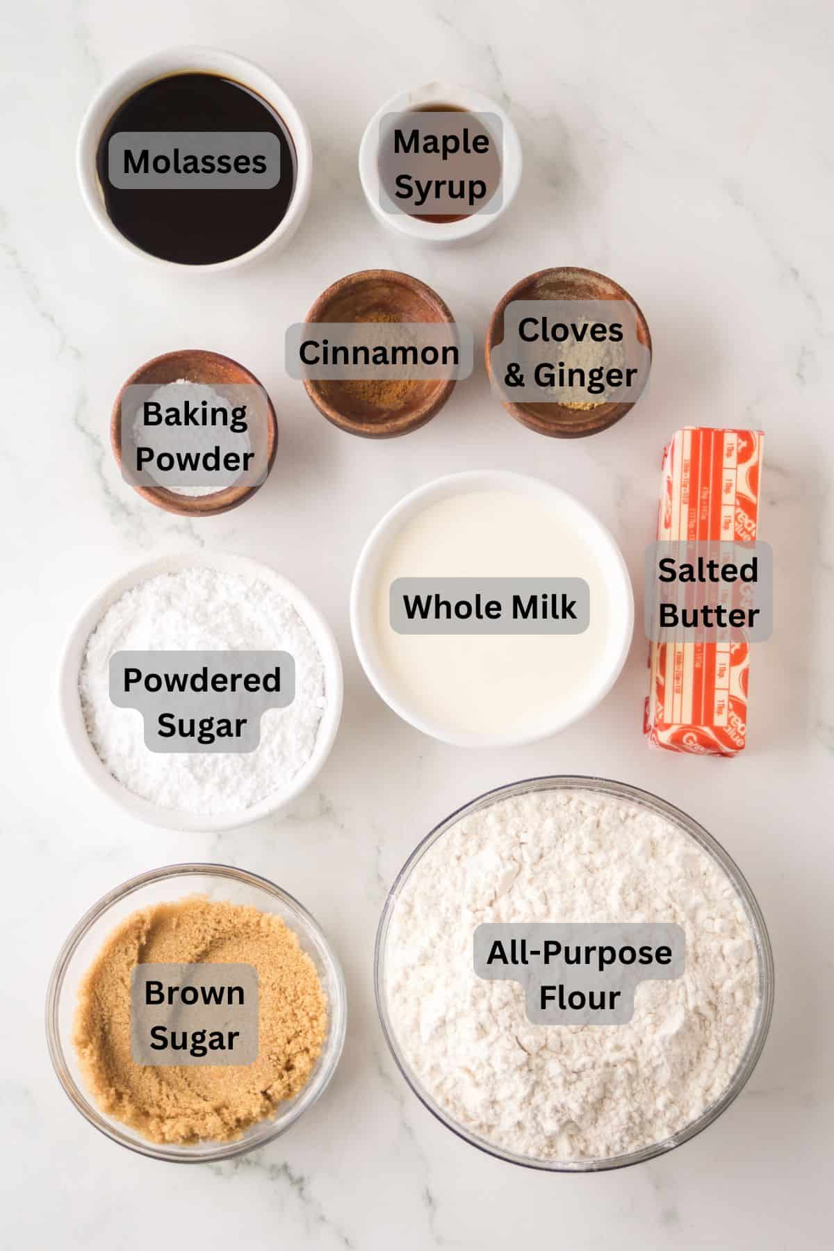 Top-down view of baking ingredients on a white surface: molasses, maple syrup, cinnamon, cloves & ginger, baking powder, powdered sugar, whole milk, brown sugar, all-purpose flour, and salted butter.