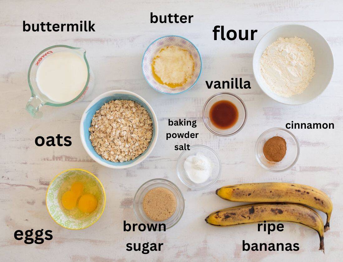 Ingredients for banana oat waffles are arranged on a surface. They include buttermilk, flour, butter, eggs, oats, ripe bananas, vanilla, cinnamon, brown sugar, baking powder, and salt, each labeled for clarity.