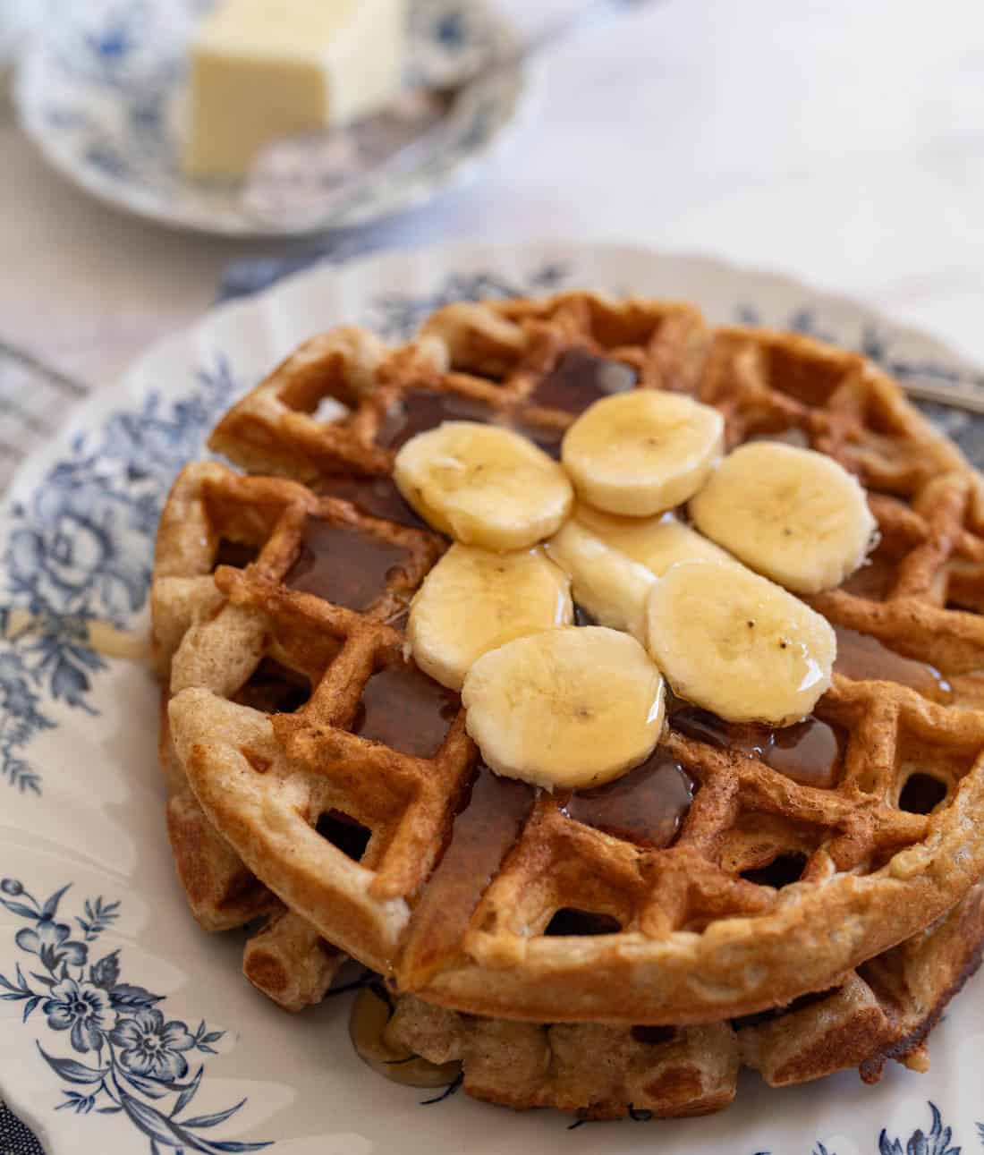 A stack of waffles topped with banana slices and syrup, served on a decorative plate. A dish with a pat of butter is blurred in the background.