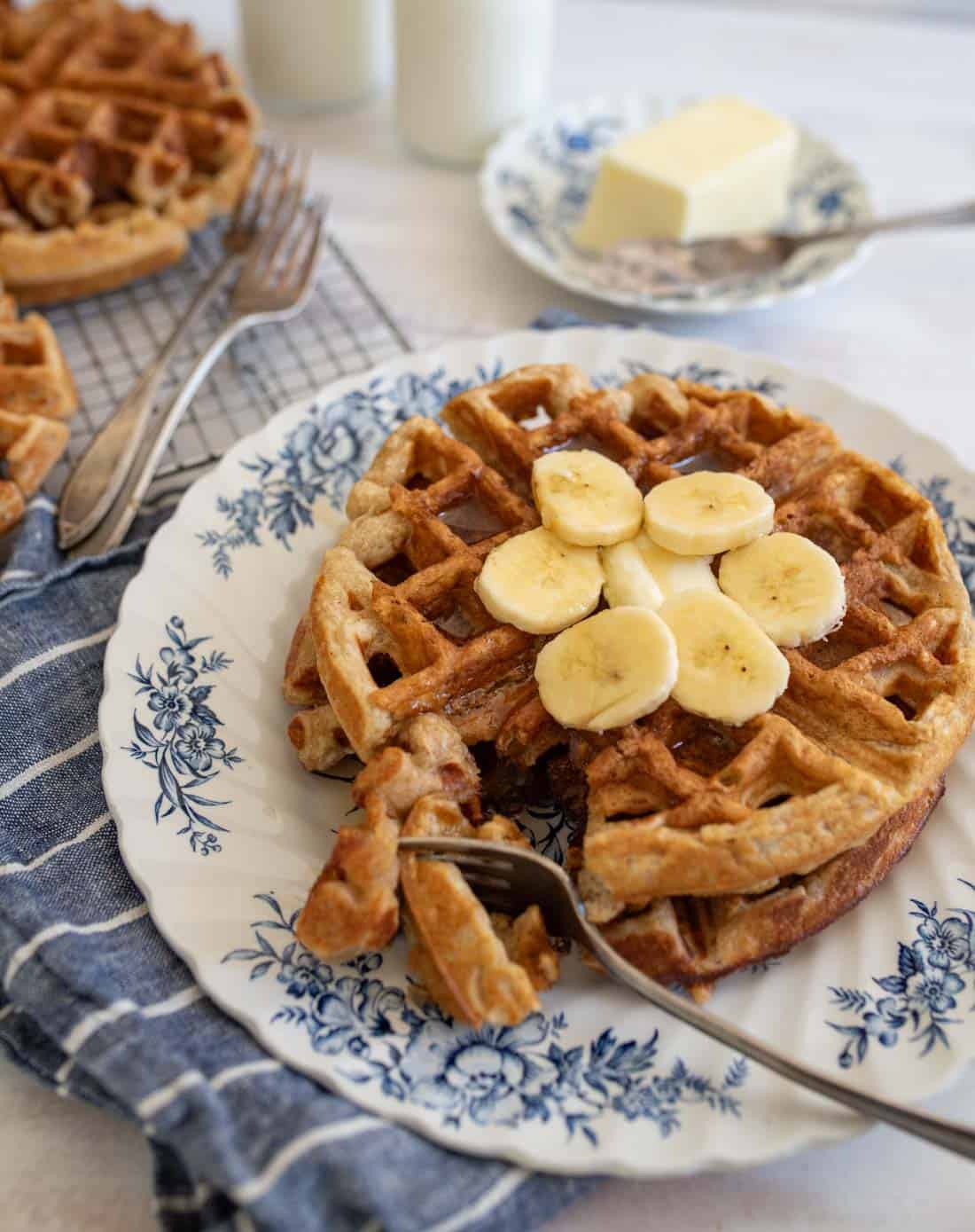 A plate with banana oatmeal waffles topped with banana slices, placed on a blue floral-patterned plate. A fork rests on the waffle. A pat of butter is on a plate in the background, with a blue cloth and other waffles nearby.