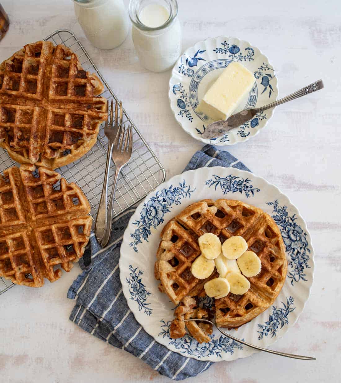 A breakfast scene with waffles on blue floral plates. One waffle is topped with banana slices. Butter is on a separate plate. Two bottles of milk are nearby. Everything is set on a blue striped cloth over a white surface.