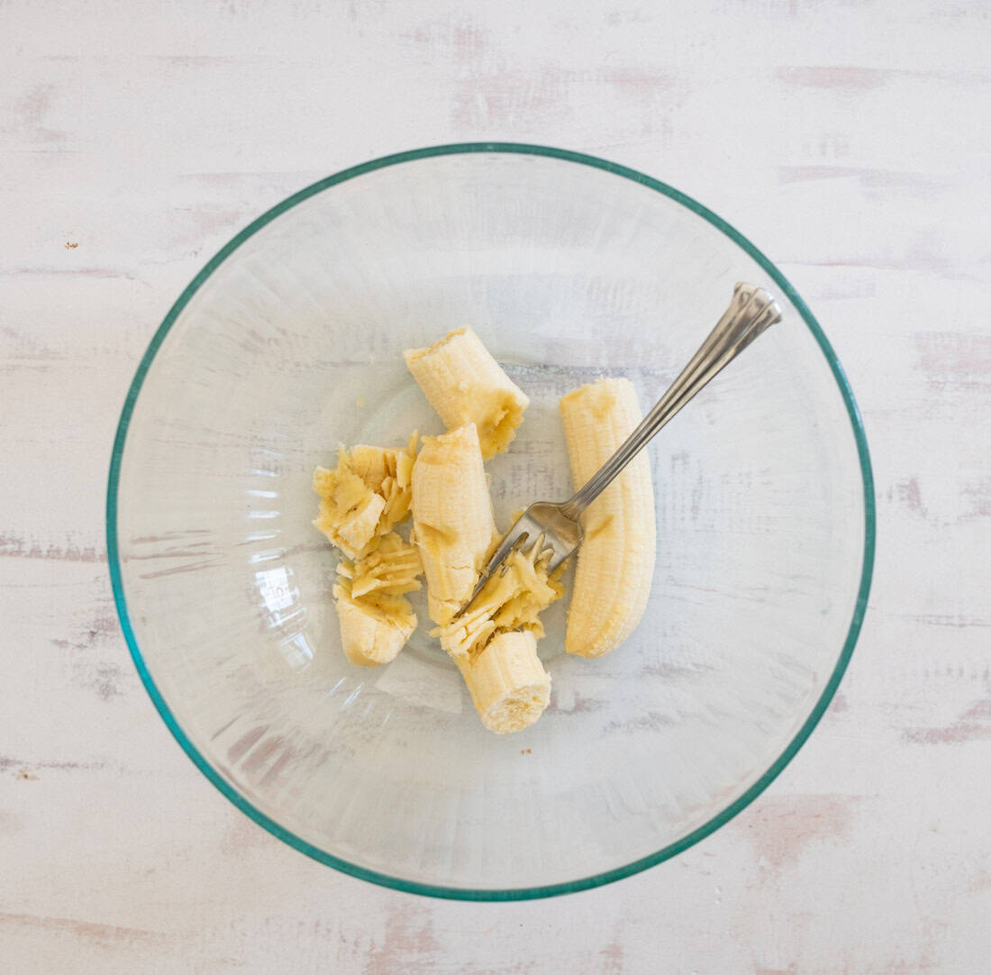 A clear glass bowl containing a peeled banana, partially mashed with a fork, sits on a light wooden surface.