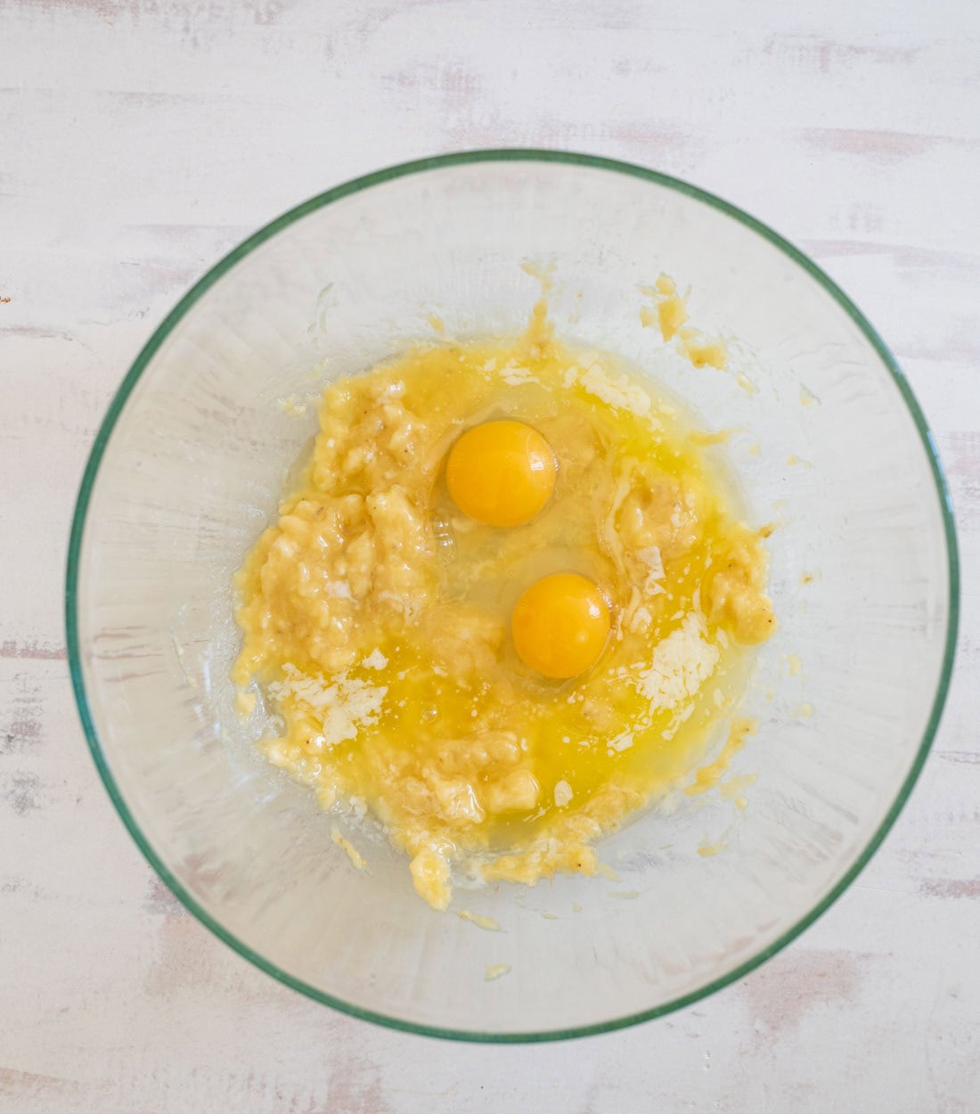 A glass bowl containing mashed bananas and two cracked eggs, ready to be mixed. The bowl is set on a light-colored wooden surface.